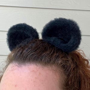 Pu&ograve; includere: Una persona con i capelli castani che indossa due scrunchies per capelli neri e pelosi nei capelli, acconciati in modo da assomigliare alle orecchie di Topolino.