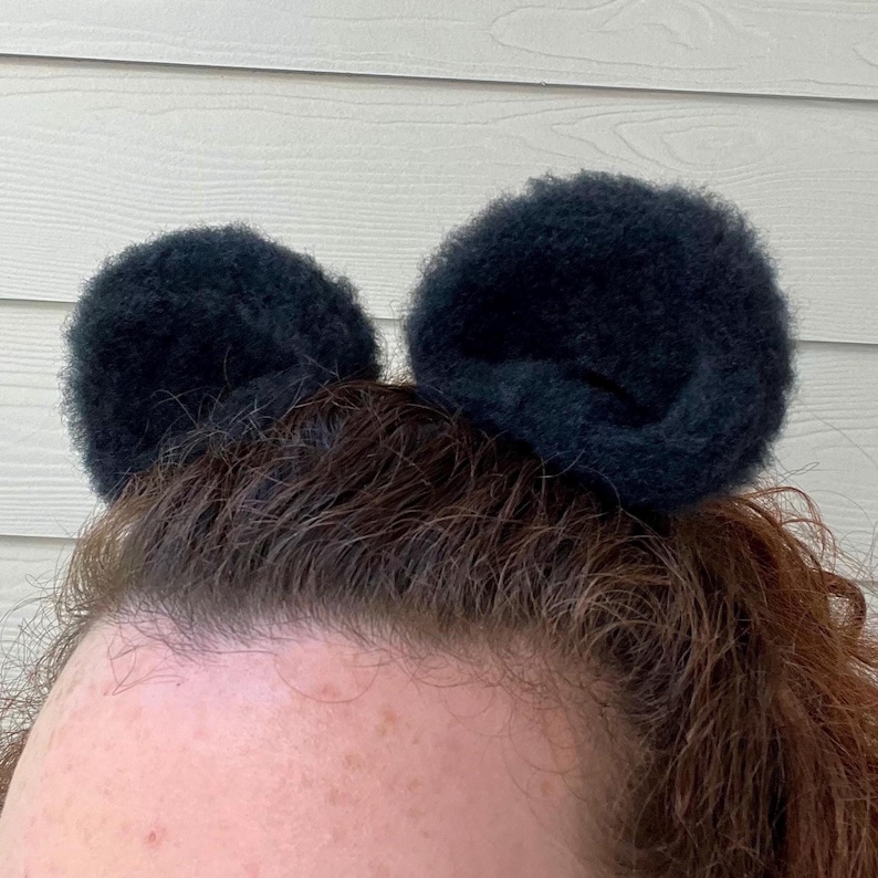 Pu&ograve; includere: Una persona con i capelli castani che indossa due scrunchies per capelli neri e pelosi nei capelli, acconciati in modo da assomigliare alle orecchie di Topolino.
