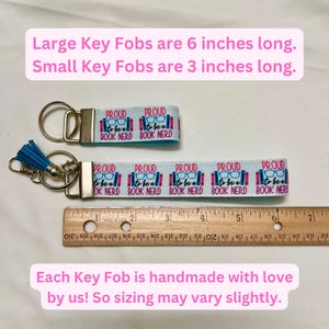 Op de afbeelding: Twee sleutelhangers met een blauw en roze lint met de tekst "Proud to be a Book Nerd". De grotere sleutelhanger meet 15 cm en de kleinere sleutelhanger meet 7,5 cm. Elke sleutelhanger is handgemaakt.