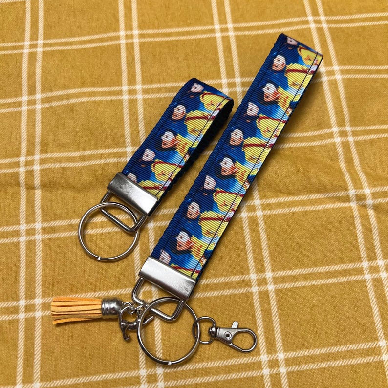 Op de afbeelding: Twee sleutelhangers met een blauw en geel gepatroneerd lint met een cartoonfiguur. De sleutelhanger aan de linkerkant heeft een zilveren ring en de sleutelhanger aan de rechterkant heeft een zilveren ring, een zilveren sluiting en een gele kwast.