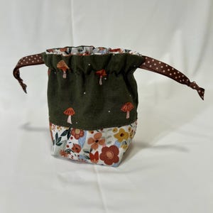 May include: A green drawstring bag with a mushroom and floral print. The bag has a brown ribbon with white polka dots.