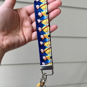 Op de afbeelding: Een blauwe en gele sleutelhanger met een herhalend patroon van een cartoonfiguur. De sleutelhanger heeft een zilveren metalen ring en een zilveren metalen sluiting.