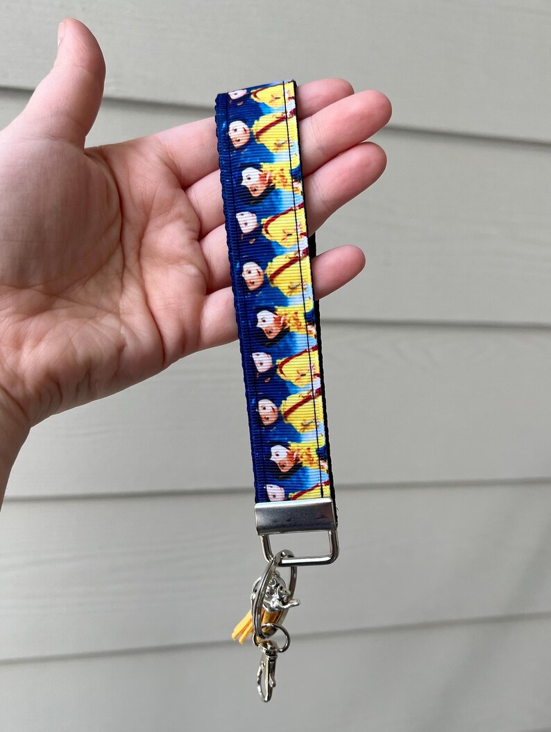 Op de afbeelding: Een blauwe en gele sleutelhanger met een herhalend patroon van een cartoonfiguur. De sleutelhanger heeft een zilveren metalen ring en een zilveren metalen sluiting.