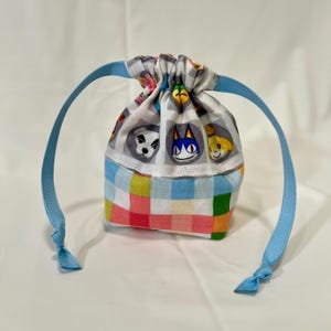 May include: A small blue and white drawstring bag with a checkered pattern. The bag features a white fabric with cartoon animal faces in circles. The bag has a blue ribbon drawstring.