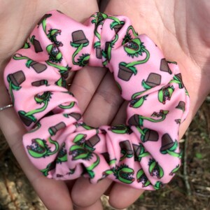 Puede incluir: Scrunchie rosa con un patrón repetido de plantas verdes de venus atrapamoscas en macetas marrones.