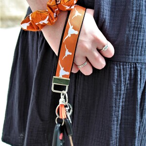 Może przedstawiać: Pomarańczowa i biała smycz z powtarzającym się wzorem longhorn, przymocowana do kluczy. Na nadgarstku noszona jest pasująca gumka do włosów. Smycz jest przymocowana do srebrnego metalowego zapięcia.