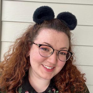 Pu&ograve; includere: Una donna con i capelli rossi ricci che indossa occhiali neri e una fascia per capelli nera con orecchie pelose. Sta sorridendo alla telecamera.