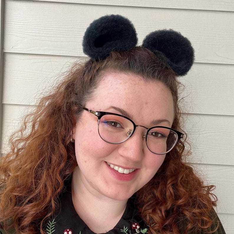 Pu&ograve; includere: Una donna con i capelli rossi ricci che indossa occhiali neri e una fascia per capelli nera con orecchie pelose. Sta sorridendo alla telecamera.