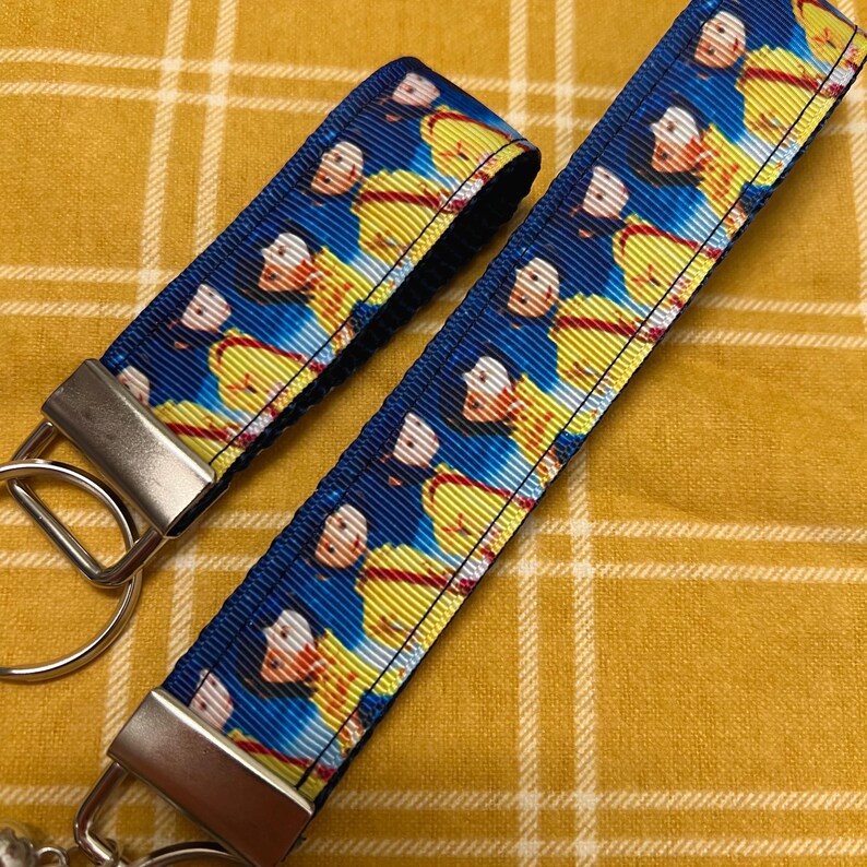 Op de afbeelding: Twee blauwe en gele sleutelhangers met een herhalend patroon van een cartoon afbeelding van Sneeuwwitje en de zeven dwergen. De sleutelhangers hebben zilveren metalen ringen.
