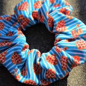 Puede incluir: Una goma para el pelo de tela con un patrón repetido de corazones de pizza sobre un fondo de rayas azules y blancas. La goma es circular y tiene un aspecto fruncido.