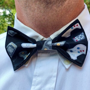 May include: A black bow tie with a white background featuring a pattern of video game controllers and game consoles.