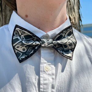 May include: A black bow tie with a white and gray pattern of video game controllers. The bow tie is tied around a white collared shirt.