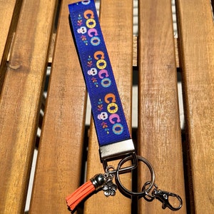 May include: A blue lanyard with the word "COCO" repeated, along with skull and floral designs. It has a silver key ring, a lobster claw clasp, and a coral tassel with a flower charm. The lanyard is on a wooden surface.