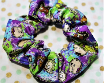 May include: A vibrant scrunchie with a repeating cartoon character design in purple, blue, green, and tan. The fabric is gathered and elasticated, forming a voluminous hair accessory.
