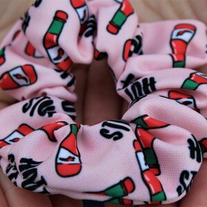 Op de afbeelding: Een roze scrunchie met een herhalend patroon van rode en groene hete sausflessen en de tekst "Hot Sauce" in zwart.