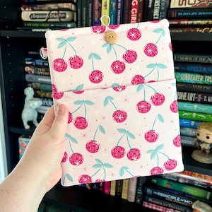 May include: A pink fabric book sleeve featuring a pattern of stylized pink cherries with disco ball-like textures and light blue stems and leaves. The sleeve has a button closure and a loop. The background is a bookshelf.
