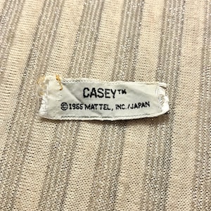 Puede incluir: Una pequeña etiqueta rectangular blanca con el nombre "CASEY" impreso en negro, junto con el texto "©1965 MATTEL, INC./JAPAN". La etiqueta está adherida a una tela beige claro acanalada.