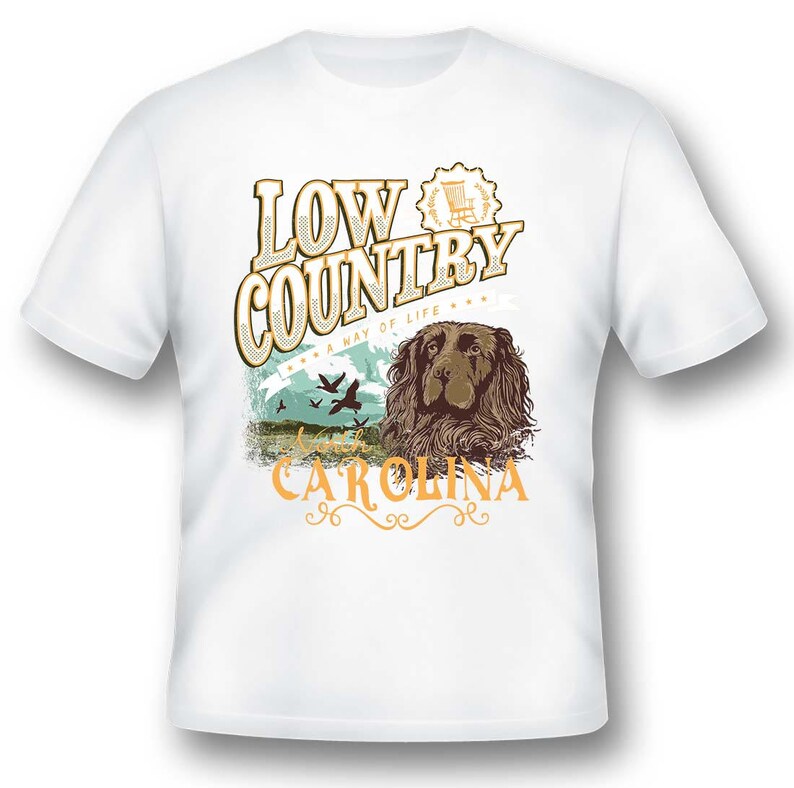 Puede incluir: Camiseta blanca con un dise&ntilde;o gr&aacute;fico marr&oacute;n y blanco que presenta un perro, p&aacute;jaros y el texto "Low Country A Way of Life South Carolina".