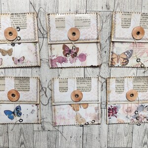 May include: Six handmade paper envelopes with butterfly and floral designs. Each envelope features a button closure and decorative stitching. The envelopes are made from vintage book pages and patterned paper, creating a unique, rustic aesthetic.