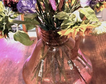 May include: A bouquet of fresh flowers in a pink glass vase. The arrangement includes sunflowers, purple roses, and other colorful blooms. The vase is pear-shaped and sits on a wooden surface.