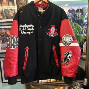 Puede incluir: Chaqueta universitaria Akademiks negra y roja con letras y parches blancos. La chaqueta presenta mangas de cuero rojas, un cuerpo negro y varios logotipos y textos bordados, incluyendo "Stadium Series 2004."