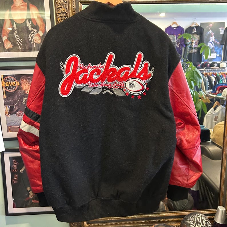 Puede incluir: Chaqueta negra con mangas de cuero rojas. La chaqueta presenta el texto "Akademiks Jackals" en letras rojas y blancas en la espalda, junto con un gr&aacute;fico de bal&oacute;n de rugby. Rayas blancas acent&uacute;an la manga izquierda.