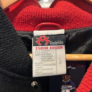 Puede incluir: Primer plano de la etiqueta de una chaqueta negra Akademiks Stadium Division. La etiqueta incluye el logotipo de la marca, la talla y la composici&oacute;n del material. La chaqueta tiene cuello y forro rojos. La manga es 100% cuero.