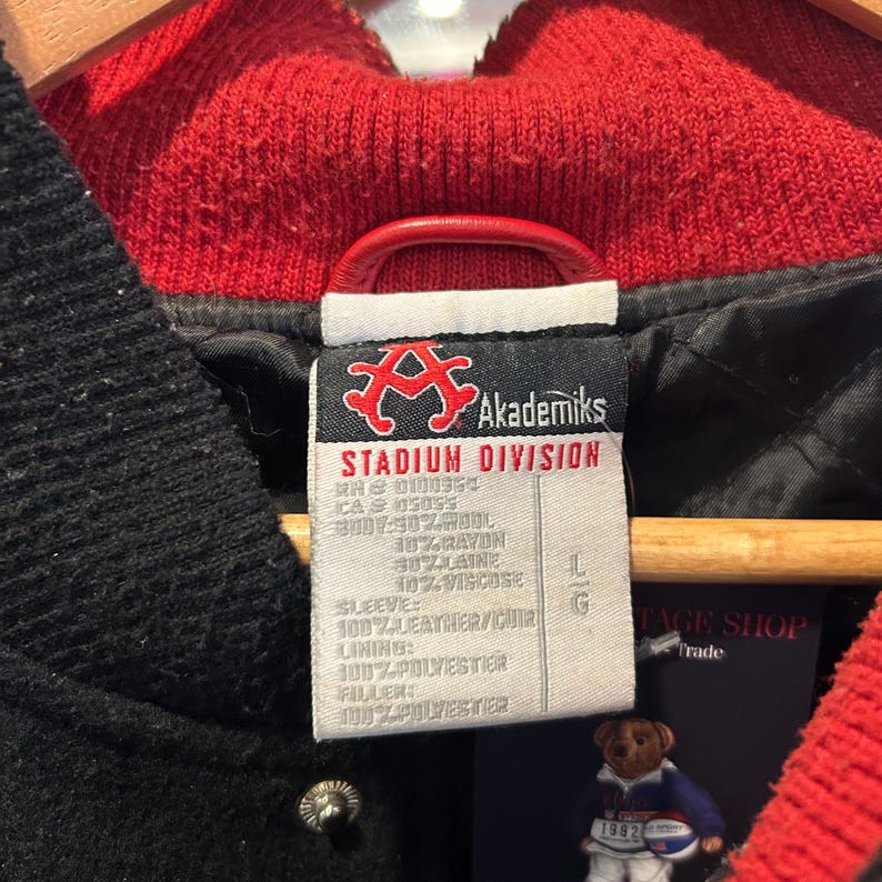 Puede incluir: Primer plano de la etiqueta de una chaqueta negra Akademiks Stadium Division. La etiqueta incluye el logotipo de la marca, la talla y la composici&oacute;n del material. La chaqueta tiene cuello y forro rojos. La manga es 100% cuero.