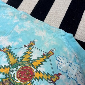 Puede incluir: Camiseta azul claro te&ntilde;ida con un dise&ntilde;o gr&aacute;fico. El dise&ntilde;o incluye un sol amarillo, una rosa roja y figuras de esqueletos. La camiseta tiene cuello redondo y mangas cortas.