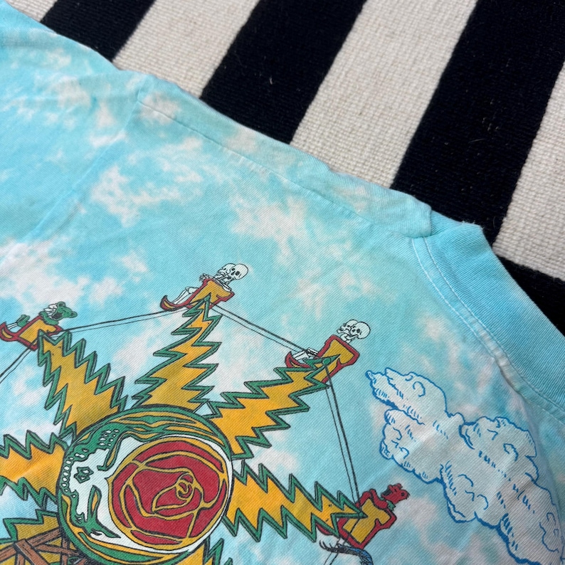 Puede incluir: Camiseta azul claro te&ntilde;ida con un dise&ntilde;o gr&aacute;fico. El dise&ntilde;o incluye un sol amarillo, una rosa roja y figuras de esqueletos. La camiseta tiene cuello redondo y mangas cortas.