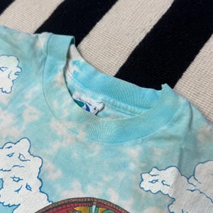 Puede incluir: Camiseta te&ntilde;ida con tinte azul claro con cuello redondo. La camiseta presenta un dise&ntilde;o de nubes y cielo con un gr&aacute;fico en la parte inferior. La etiqueta dice "100% Algod&oacute;n" y "L".