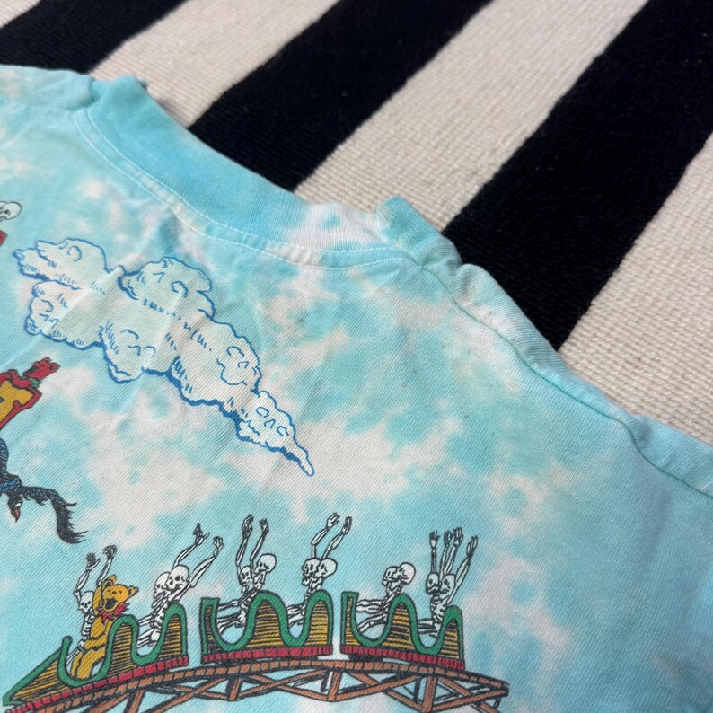 Puede incluir: Camiseta tie-dye azul claro con una escena de monta&ntilde;a rusa con jinetes esquel&eacute;ticos y un oso de dibujos animados. El dise&ntilde;o incluye nubes y una pista de monta&ntilde;a rusa de madera. La camiseta tiene cuello redondo y mangas cortas.