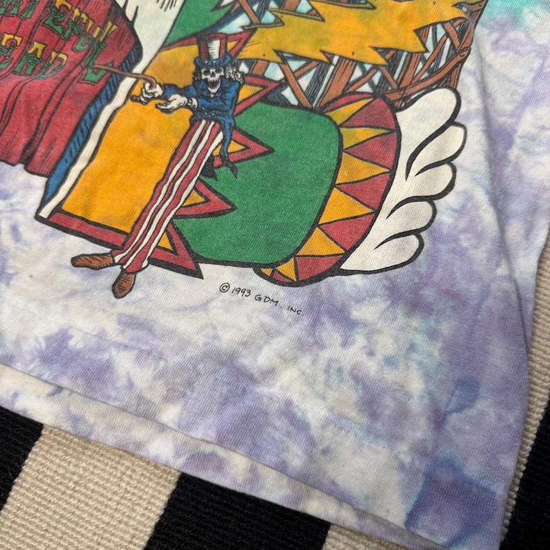 Puede incluir: Camiseta te&ntilde;ida con un dise&ntilde;o gr&aacute;fico colorido. El dise&ntilde;o incluye una figura de esqueleto con abrigo azul y sombrero de copa, sosteniendo una batuta. El gr&aacute;fico se establece sobre un fondo de elementos rojos, amarillos, verdes y blancos. La camiseta tiene un patr&oacute;n te&ntilde;ido en tonos morados y blancos.