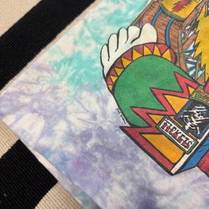 Puede incluir: Una camiseta te&ntilde;ida con un dise&ntilde;o gr&aacute;fico colorido. El gr&aacute;fico presenta una figura nativa americana con un tocado, un dise&ntilde;o verde y rojo, y la palabra "TICKETS" en un recuadro. La camiseta tiene un patr&oacute;n de te&ntilde;ido anudado azul, morado y blanco.