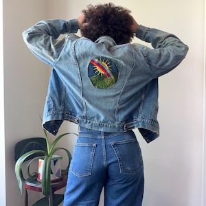 May include: Light blue denim jacket with a round embroidered patch on the back featuring a rainbow, sun, and green hills. The jacket has a classic design with button closures and pockets. The model is wearing blue jeans.