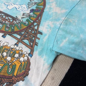 Puede incluir: Camiseta te&ntilde;ida con tinte azul claro con un gr&aacute;fico colorido de una monta&ntilde;a rusa con jinetes esquel&eacute;ticos. La monta&ntilde;a rusa es marr&oacute;n y amarilla, con esqueletos blancos. El cielo es azul claro con nubes blancas.