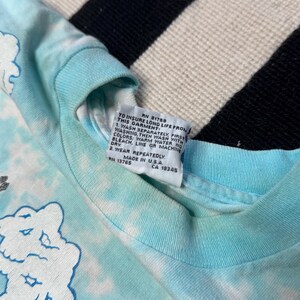 Puede incluir: Una camiseta azul claro te&ntilde;ida con gr&aacute;ficos de nubes blancas. La camiseta tiene una etiqueta con instrucciones de cuidado y el texto "MADE IN U.S.A."