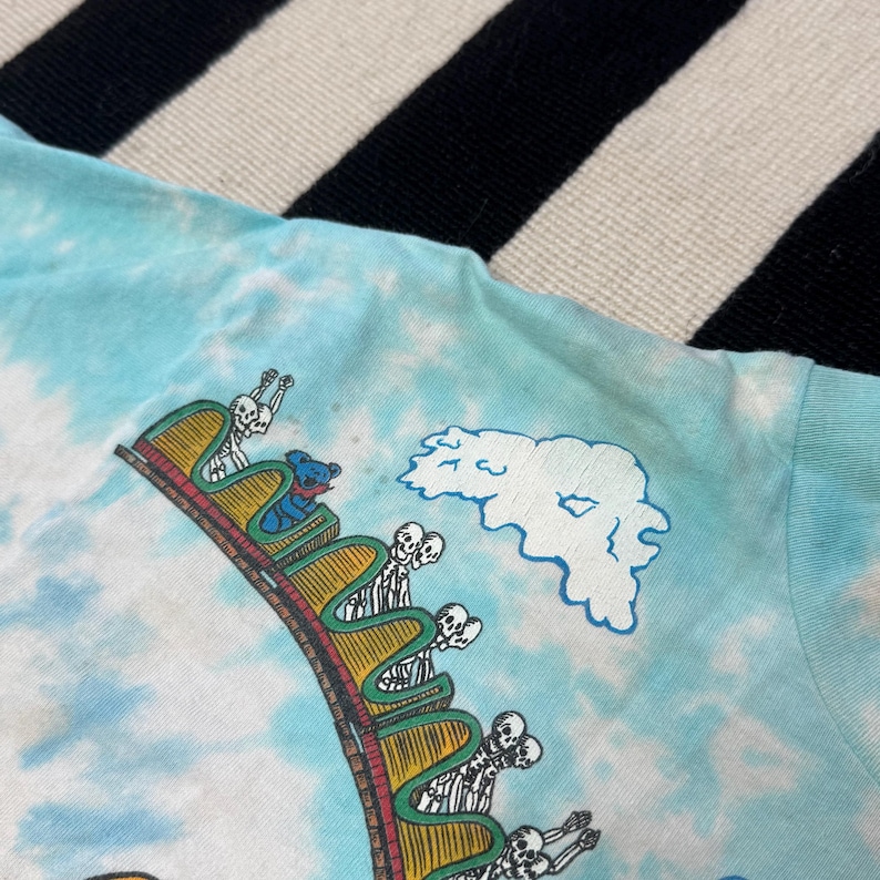 Puede incluir: Camiseta azul claro te&ntilde;ida con un dise&ntilde;o de monta&ntilde;a rusa con jinetes esquel&eacute;ticos y un oso azul. El dise&ntilde;o incluye un gr&aacute;fico de nube. Una prenda &uacute;nica y colorida.