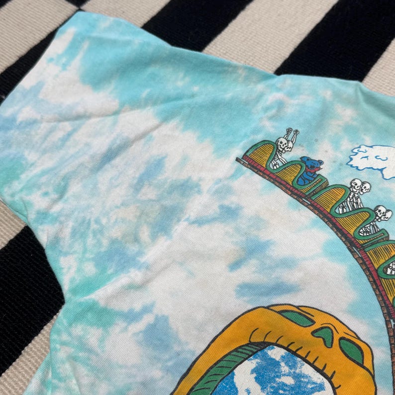 Puede incluir: Una camiseta te&ntilde;ida con un patr&oacute;n de nubes azules y blancas. La camiseta presenta un gr&aacute;fico colorido de una monta&ntilde;a rusa con jinetes esquel&eacute;ticos y un oso. La monta&ntilde;a rusa est&aacute; sobre un cielo azul.