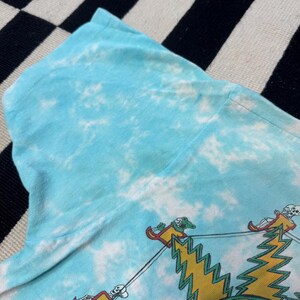 Puede incluir: Camiseta azul claro te&ntilde;ida con un dise&ntilde;o de esqueletos en una atracci&oacute;n de feria. El dise&ntilde;o incluye rayos amarillos y coches de feria rojos y amarillos. La camiseta tiene cuello redondo y mangas cortas.