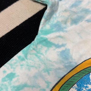 Puede incluir: Primer plano de una manga de camiseta te&ntilde;ida con tinte anudado. La tela presenta una mezcla de colores blanco, azul claro y verde azulado. La manga est&aacute; colocada sobre una superficie a rayas en blanco y negro.