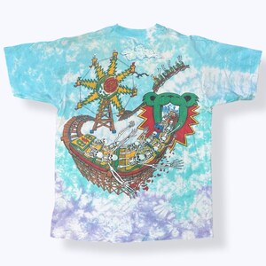 Puede incluir: Camiseta te&ntilde;ida con un dise&ntilde;o gr&aacute;fico colorido de una monta&ntilde;a rusa con esqueletos, una noria y un emblema de oso. La camiseta tiene un patr&oacute;n de te&ntilde;ido anudado azul, morado y blanco.