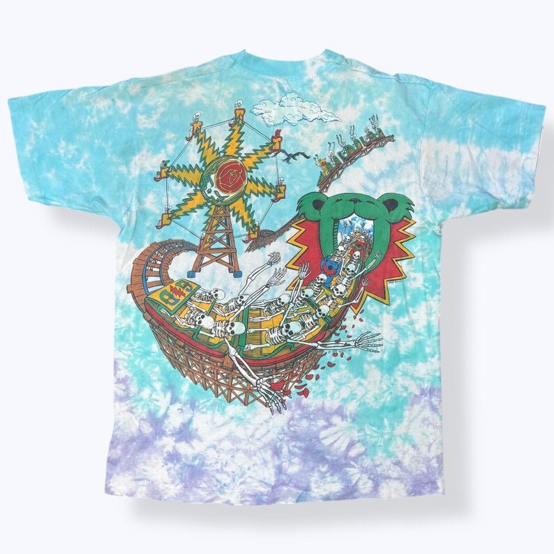 Puede incluir: Camiseta te&ntilde;ida con un dise&ntilde;o gr&aacute;fico colorido de una monta&ntilde;a rusa con esqueletos, una noria y un emblema de oso. La camiseta tiene un patr&oacute;n de te&ntilde;ido anudado azul, morado y blanco.