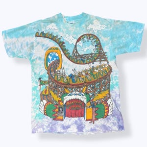 Puede incluir: Camiseta te&ntilde;ida con un dise&ntilde;o de parque de atracciones colorido. El dise&ntilde;o incluye una monta&ntilde;a rusa con esqueletos, nubes y las palabras "Grateful Dead". La camiseta tiene un patr&oacute;n de te&ntilde;ido anudado azul, morado y blanco.