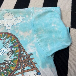 Puede incluir: Una camiseta te&ntilde;ida con tinte azul claro con un gr&aacute;fico colorido de una noria con figuras de esqueletos. El dise&ntilde;o incluye una estructura de monta&ntilde;a rusa de madera y formas de nubes blancas. Se ve la manga de la camiseta.