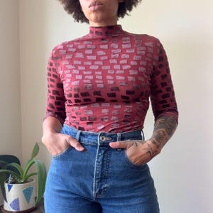 May include: A burgundy mock neck top with a brick pattern design. The top has three-quarter sleeves and is paired with blue denim jeans. The jeans have a classic five-pocket design.