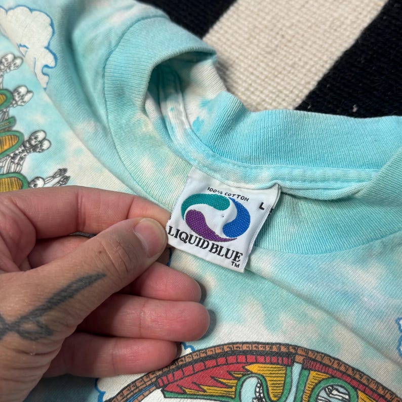 Puede incluir: Camiseta te&ntilde;ida anudada azul claro con una etiqueta Liquid Blue. La etiqueta es blanca con un logotipo azul y morado y las palabras "LIQUID BLUE" en negro. La camiseta es 100% algod&oacute;n y talla L.