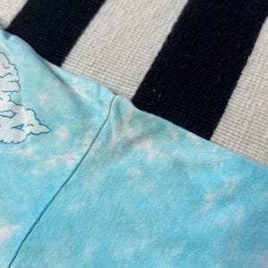 Puede incluir: Primer plano de una manga de camiseta azul claro te&ntilde;ida con tinte anudado. La camisa tiene un dise&ntilde;o de nubes en blanco y azul claro. La manga est&aacute; sobre una superficie a rayas blancas y negras.