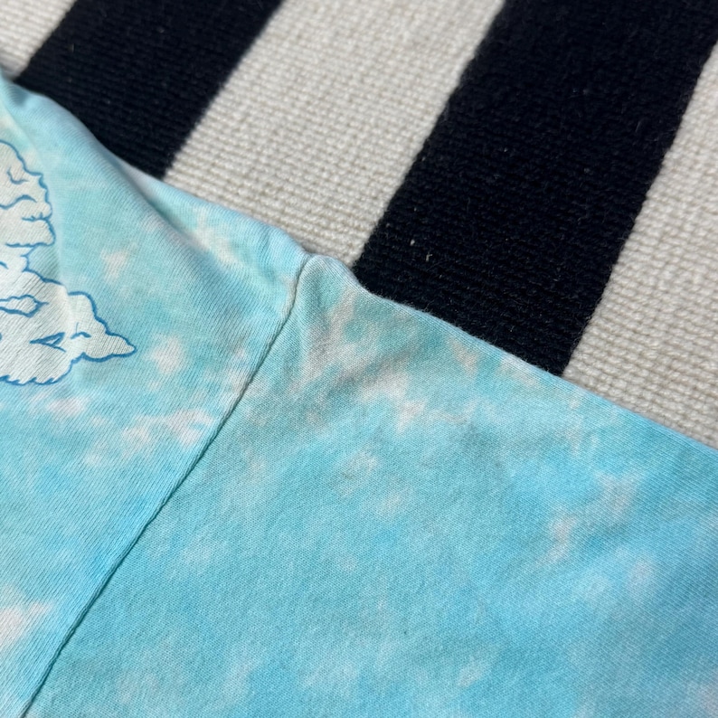 Puede incluir: Primer plano de una manga de camiseta azul claro te&ntilde;ida con tinte anudado. La camisa tiene un dise&ntilde;o de nubes en blanco y azul claro. La manga est&aacute; sobre una superficie a rayas blancas y negras.