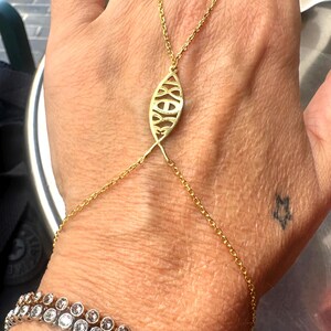 May include: A gold-colored hand chain with a decorative oval centerpiece and delicate chain accents. A diamond bracelet is also visible. The jewelry is worn on a hand with a star tattoo.
